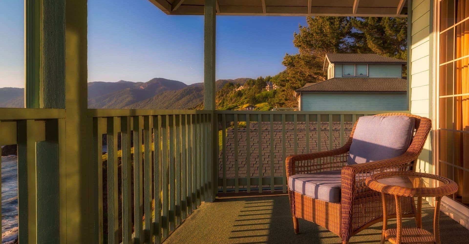 A balcony with a wicker chair and a view of mountains and houses.