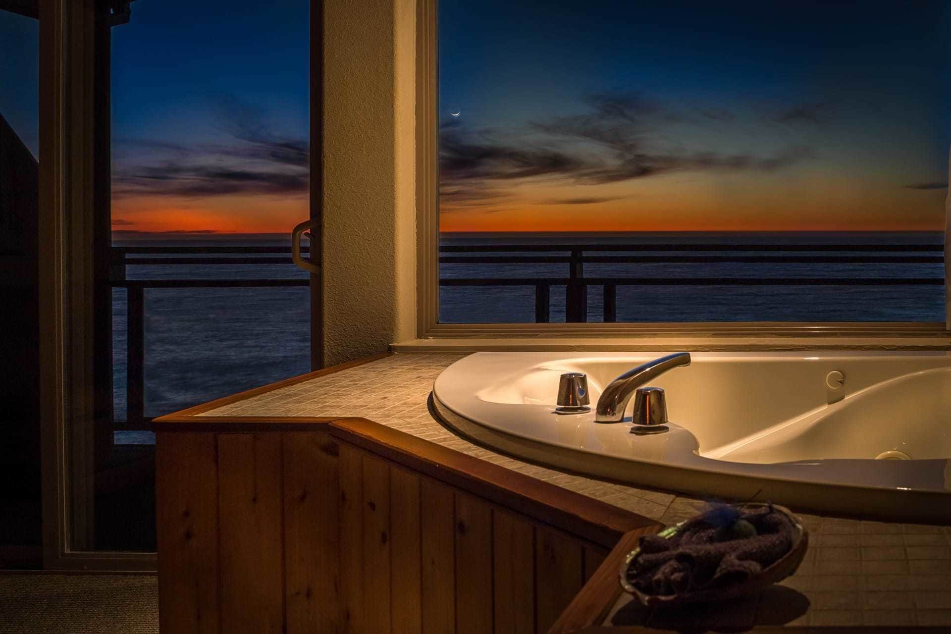 A bathroom with a large bathtub, a wooden vanity, and a view of the ocean at sunset.