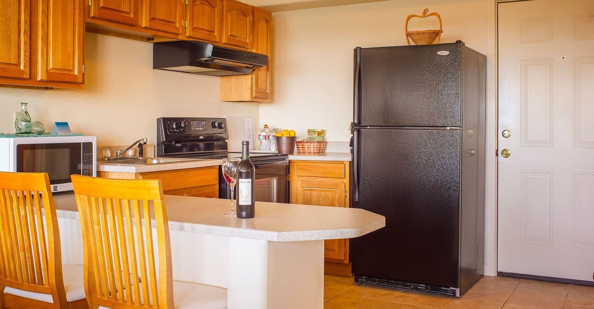 A kitchen with wooden cabinets, a black refrigerator, a microwave, and a wine bottle on the counter.