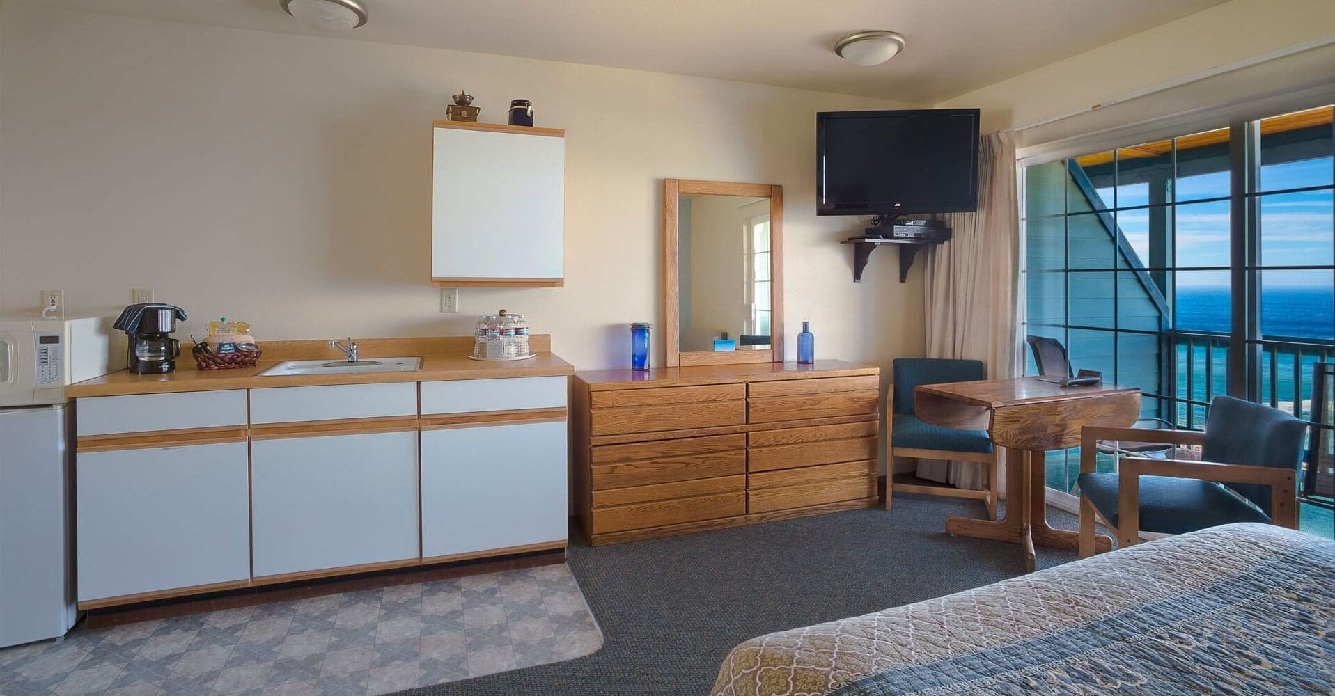 Habitación de hotel con kitchenette, un espejo grande, un televisor de pantalla plana, un escritorio con silla y vista al océano.