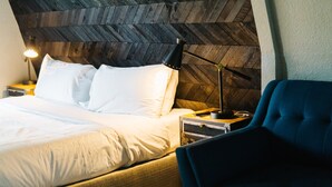 A neatly made bed with white linens, a black lamp on a bedside table, and a blue chair in a room with a herringbone-patterned wooden headboard. - Vintage Charm: Walk to CSU & Old Town (Fort Collins)