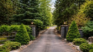 Un chemin pavé, bordé de murs en pierre et d’une végétation luxuriante, mène à une entrée fermée par un portail.