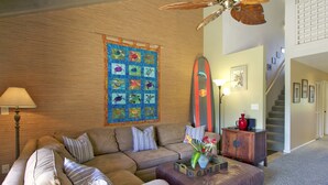 A living room with a wooden wall, a large quilt, a surfboard, a sofa, a coffee table, and a staircase. - Quiet, Private Paradise M208 steps to the tropical pool and tennis court (Kihei)