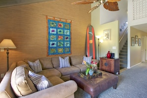 A living room with a wooden wall, a large quilt, a surfboard, a sofa, a coffee table, and a staircase.