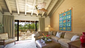A spacious living room with a large window, a ceiling fan, a wooden ceiling, and a colorful quilt on the wall. - Quiet, Private Paradise M208 steps to the tropical pool and tennis court (Kihei)