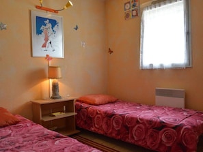 Quarto com duas camas de solteiro, mesa de cabeceira, abajur, janela com cortinas e relógio na parede.