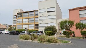 Un immeuble à plusieurs étages doté de balcons, un parking avec plusieurs voitures et un petit espace paysager agrémenté d’arbustes et d’un arbre.