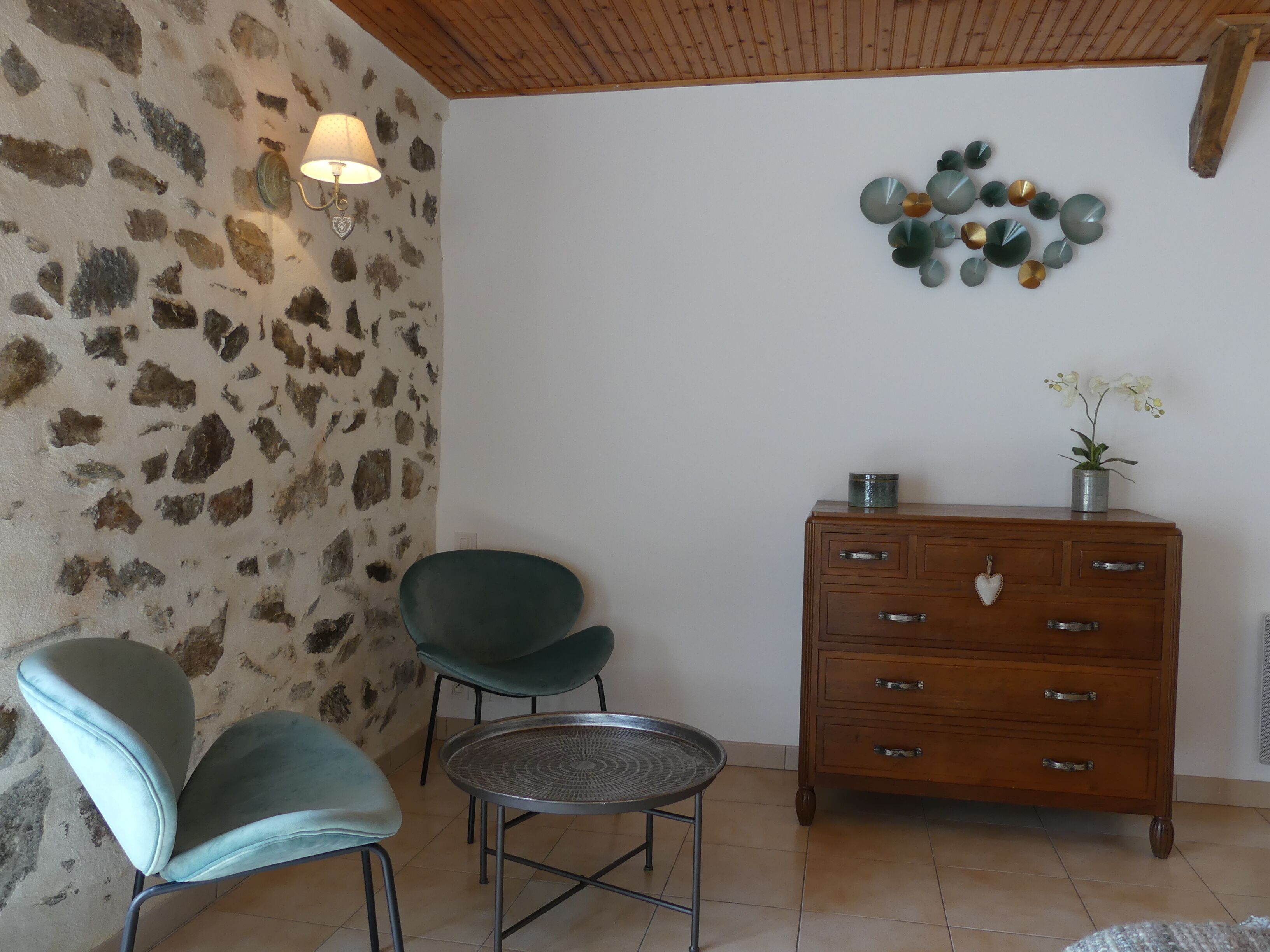 Una habitación con pared de piedra, dos sillas, una mesita redonda pequeña, un aparador de madera y una lámpara fijada en la pared.