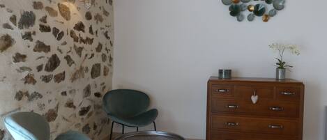 Una habitación con pared de piedra, dos sillas, una mesita redonda pequeña, un aparador de madera y una lámpara fijada en la pared.