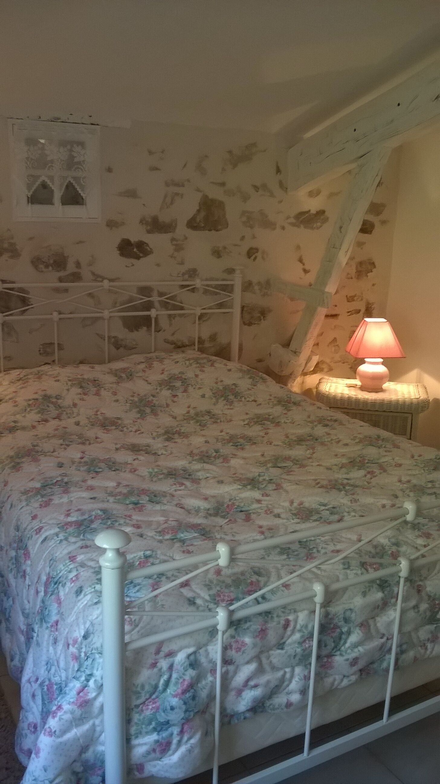 A bedroom with a floral bedspread, a white metal bed frame, a nightstand with a lamp, and a stone-patterned wall.
