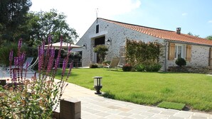 Un bâtiment en pierre avec un toit en tuiles, une pelouse dallée et un petit jardin agrémenté de fleurs violettes.