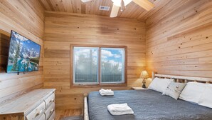 A wooden cabin room with a bed, a dresser, a window with blinds, and a painting on the wall.