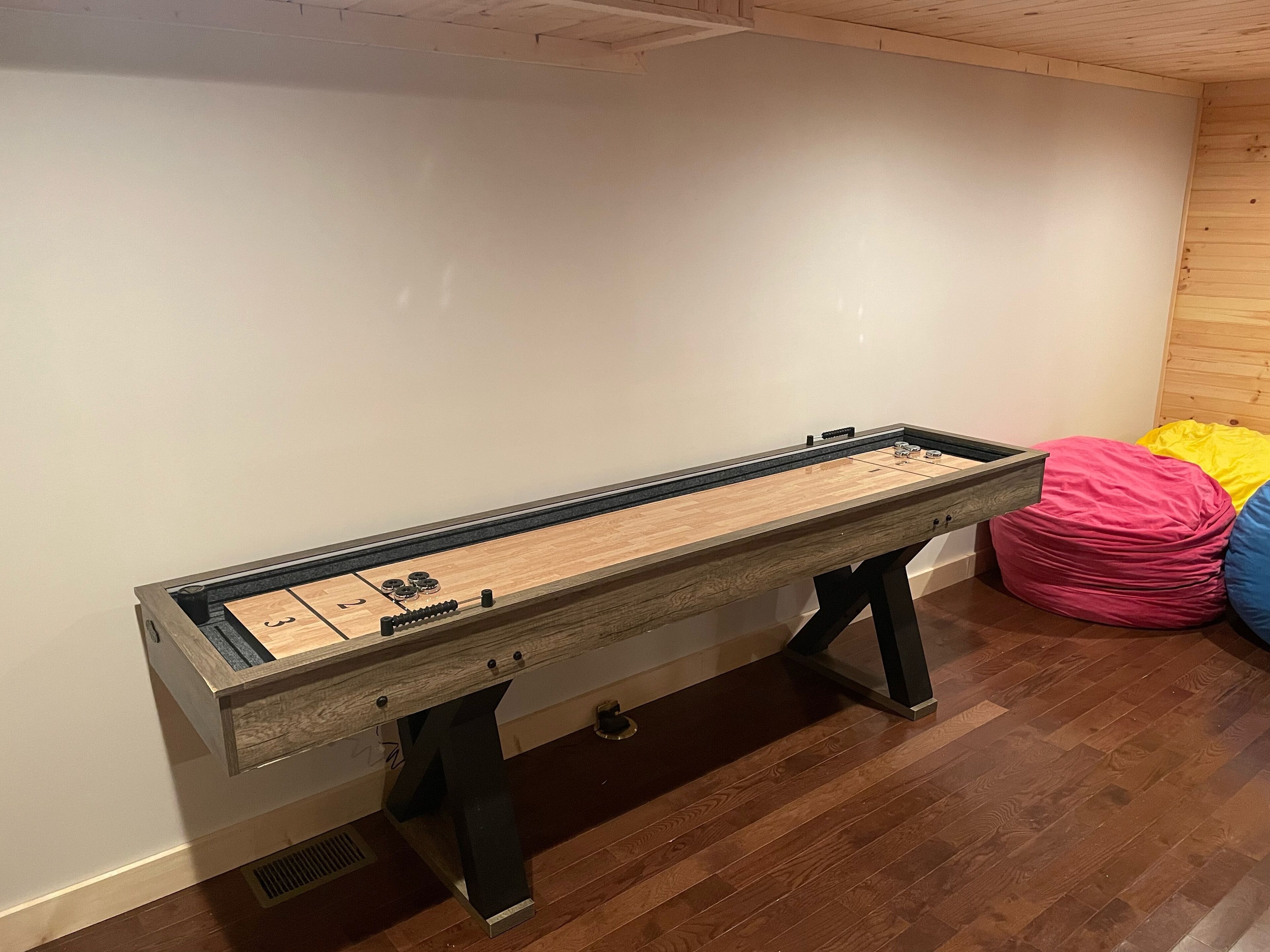 Uma mesa de jogo de shuffleboard com acabamento em madeira, colocada em frente a uma parede branca.