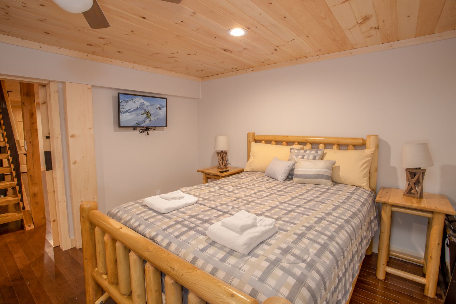 Una cama con estructura de madera y colcha a cuadros, dos mesitas de noche con lámparas, un televisor montado y una escalera de madera que conduce a un nivel superior.