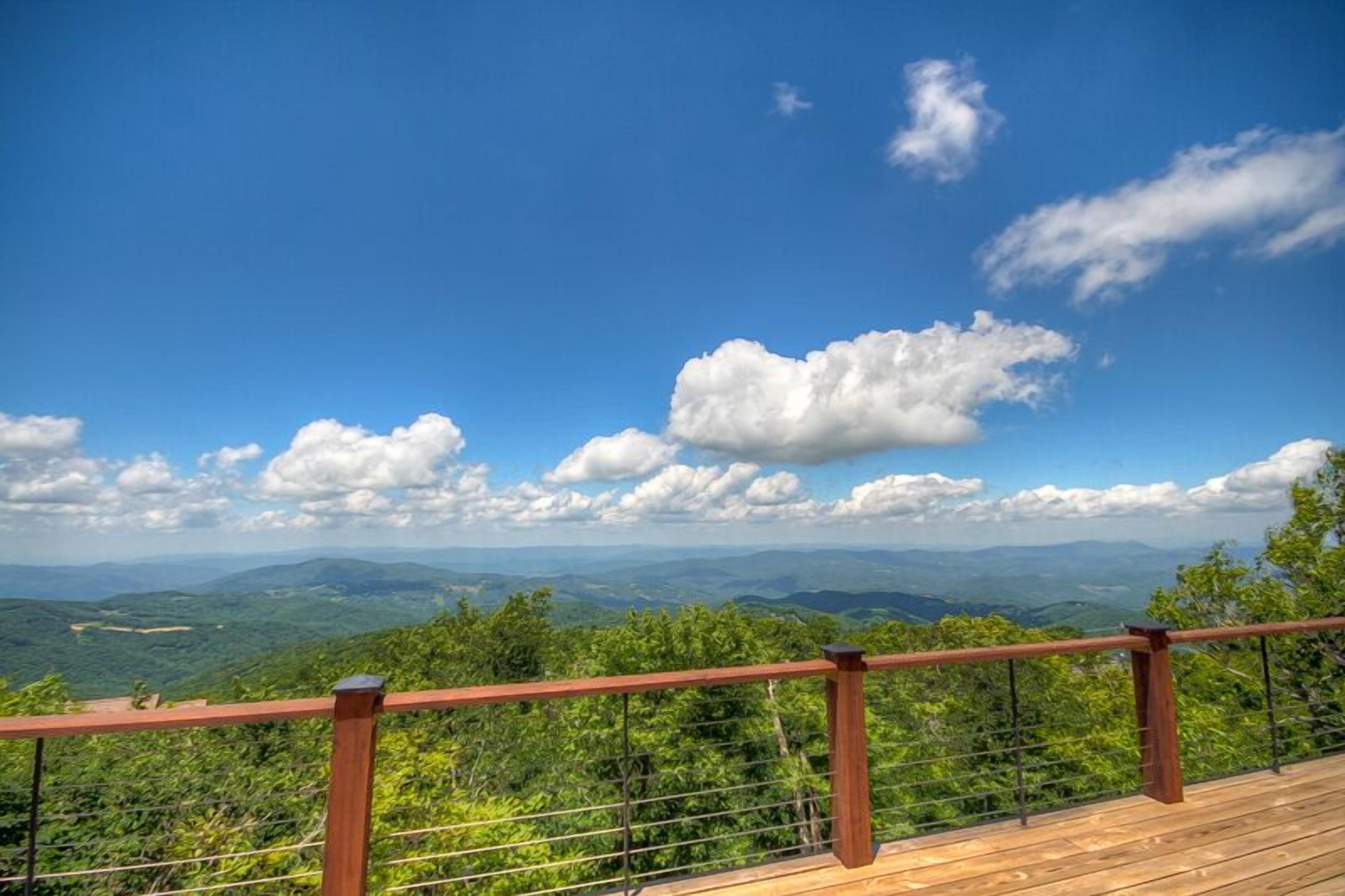 A wooden deck with a metal railing overlooking a mountainous landscape with lush greenery and a clear blue sky.