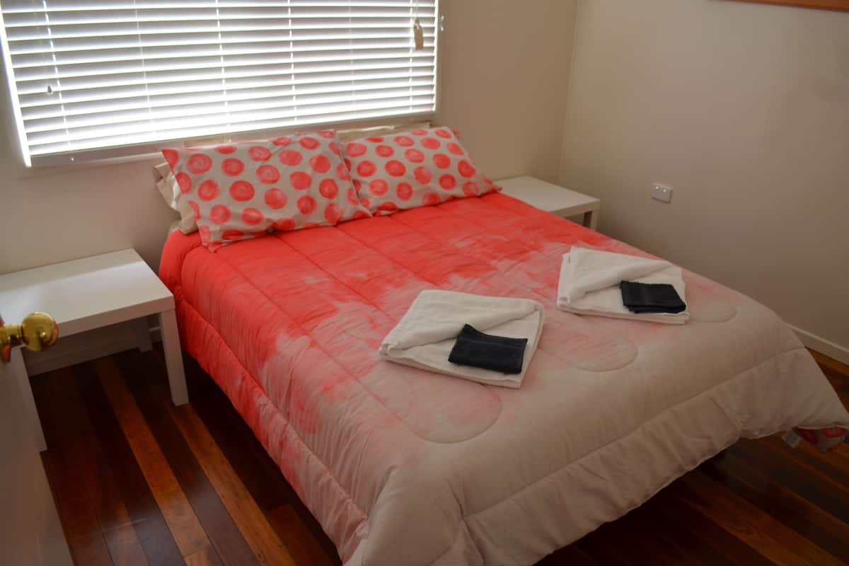 Una cama bien hecha con una colcha roja y blanca, dos almohadas con estampado de lunares y dos toallas dobladas sobre la cama.