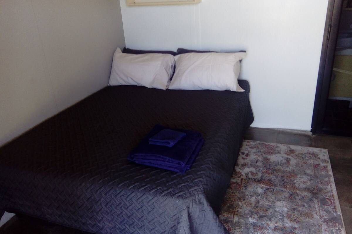Una cama con una colcha oscura y dos almohadas, una toalla azul doblada y un tapete estampado con motivos florales.