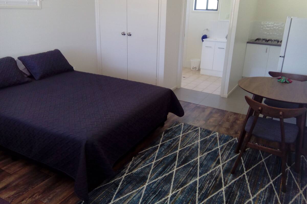Un dormitorio con cama, una mesita y una silla. Hay una puerta que da a un baño y una cocina.