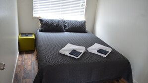 A neatly made bed with a dark quilt and two pillows, a green suitcase, and a window with blinds.