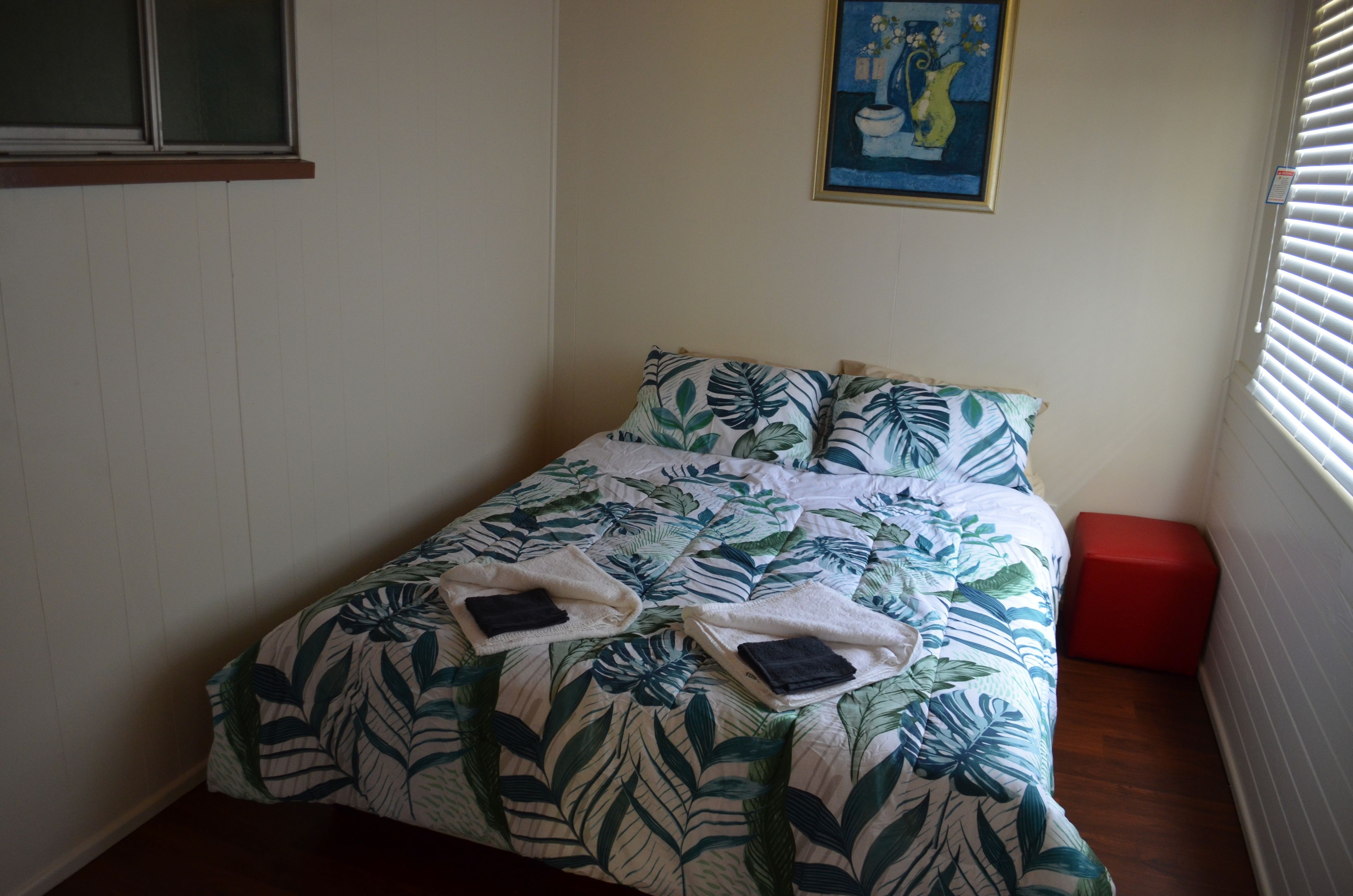 Un dormitorio con una cama con colcha de hojas tropicales, dos almohadas y dos toallas dobladas. Un reposapiés rojo está apoyado contra la pared, y un cuadro cuelga sobre la cama.