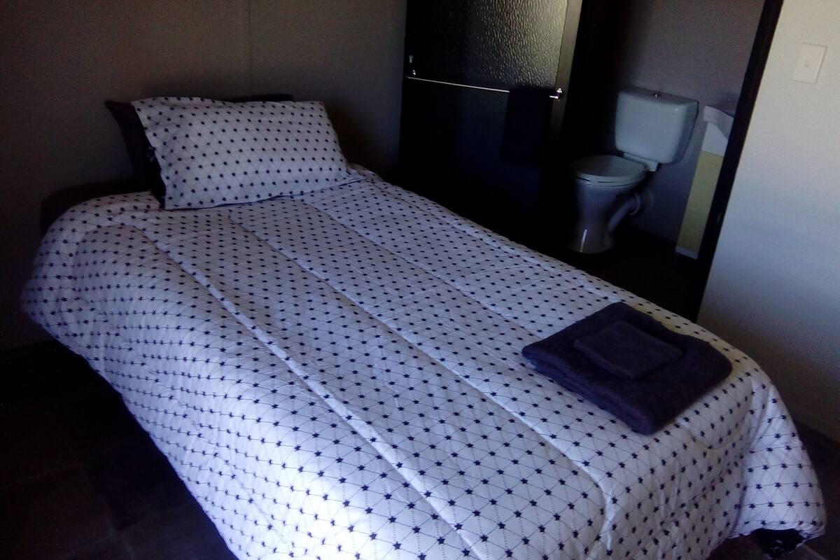 Una cama individual con una colcha blanca con estampado de estrellas, una almohada de color oscuro y una toalla doblada de color oscuro sobre la cama.