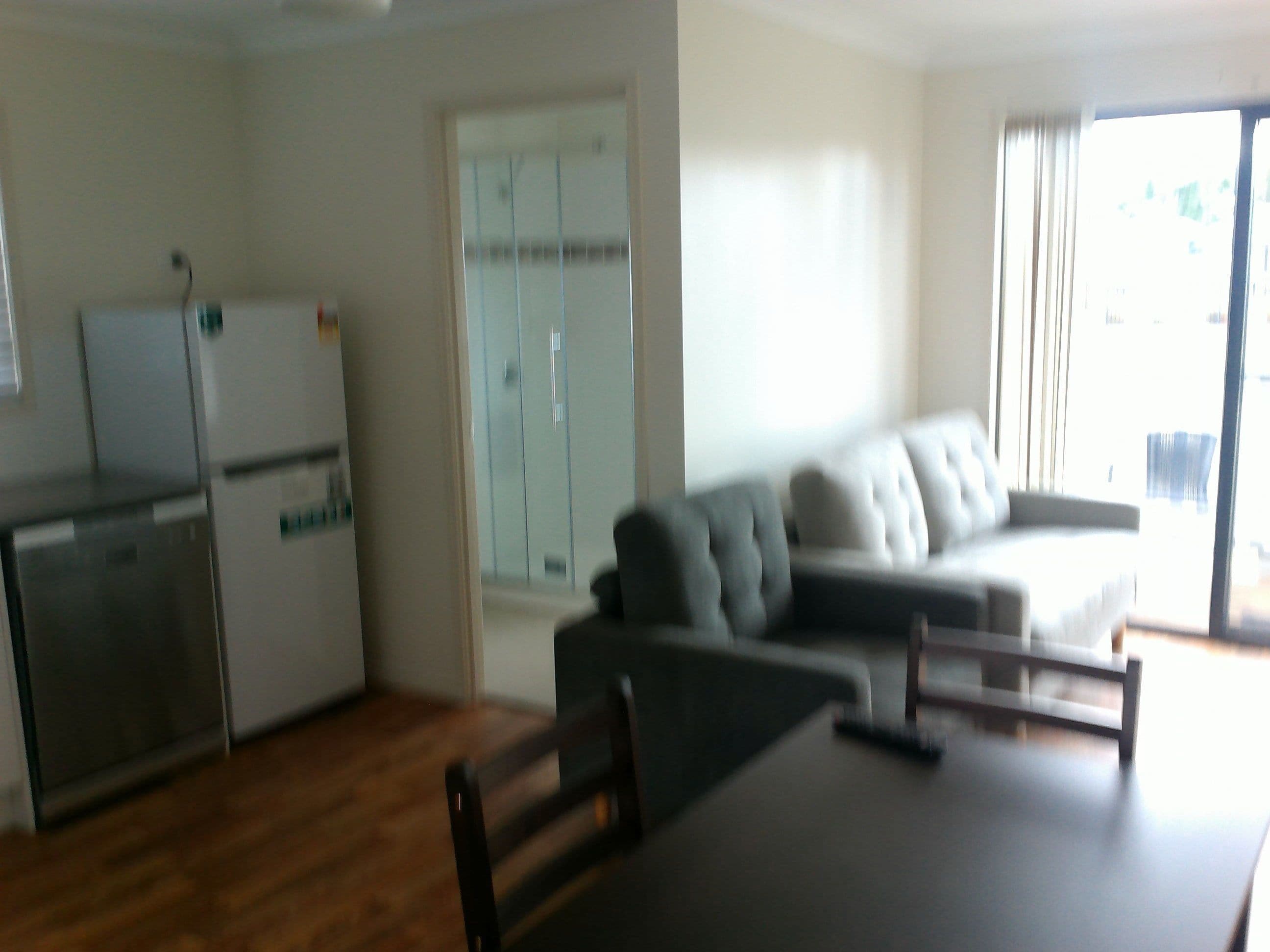 A modern living room with a grey sofa, a dining table, and a kitchen area with a refrigerator and cabinets.