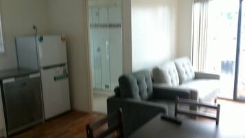 A modern living room with a grey sofa, a dining table, and a kitchen area with a refrigerator and cabinets.