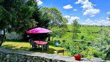 A stone wall with a table and chairs under a pink umbrella, surrounded by greenery and a view of rolling hills.