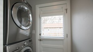 A laundry room with a front-loading washer and dryer, a white door with a window, and a view of a building outside.