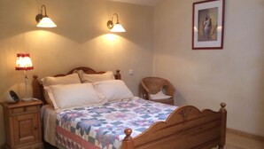 A wooden bed with a quilt, two wall-mounted lamps, a nightstand with a lamp, a wicker chair, and a framed picture on the wall. - Cottage rental Les tilleuls stone house in Perigord Noir Sarlat (Castels)