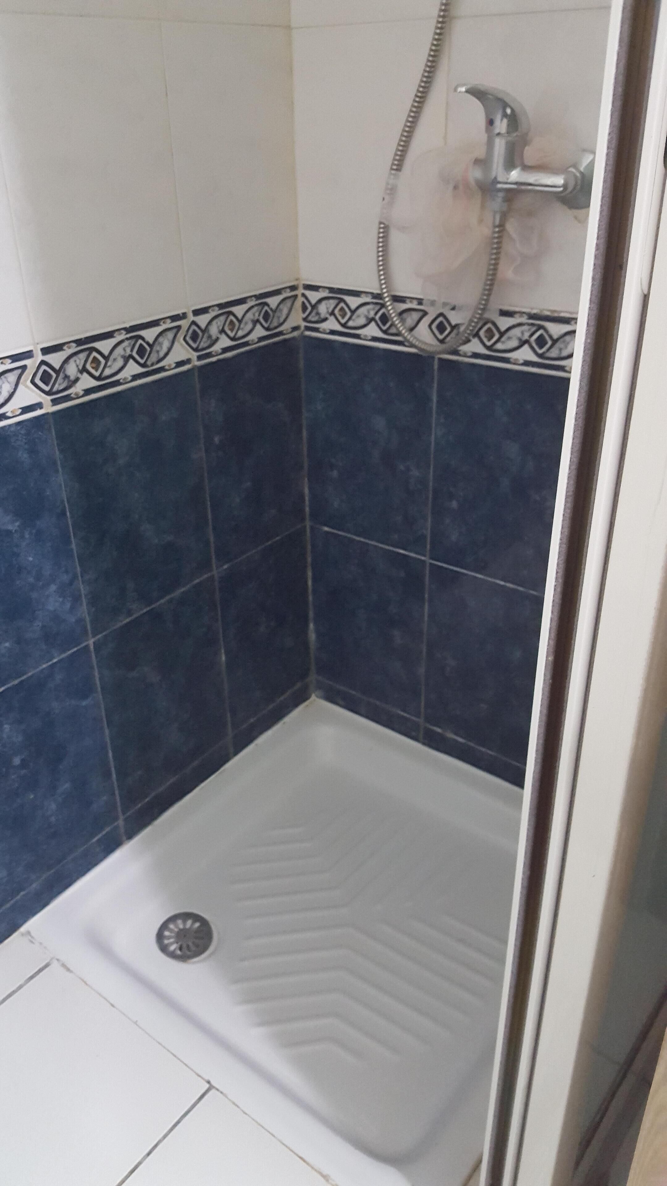 Zona de ducha con suelo blanco, cabezal de ducha y desagüe. Las paredes están revestidas con azulejos azules y blancos.