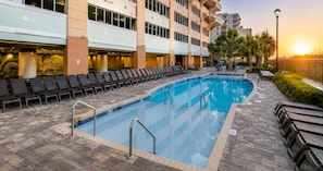 A swimming pool with lounge chairs, a hotel building, and a sunset in the background.