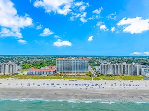 A coastal cityscape with a prominent hotel featuring multiple floors and balconies, situated near a beach with numerous umbrellas and people. - Blue Haven · Direct Oceanfront 3BR, Sleeps 10, High Floor (North Myrtle Beach)