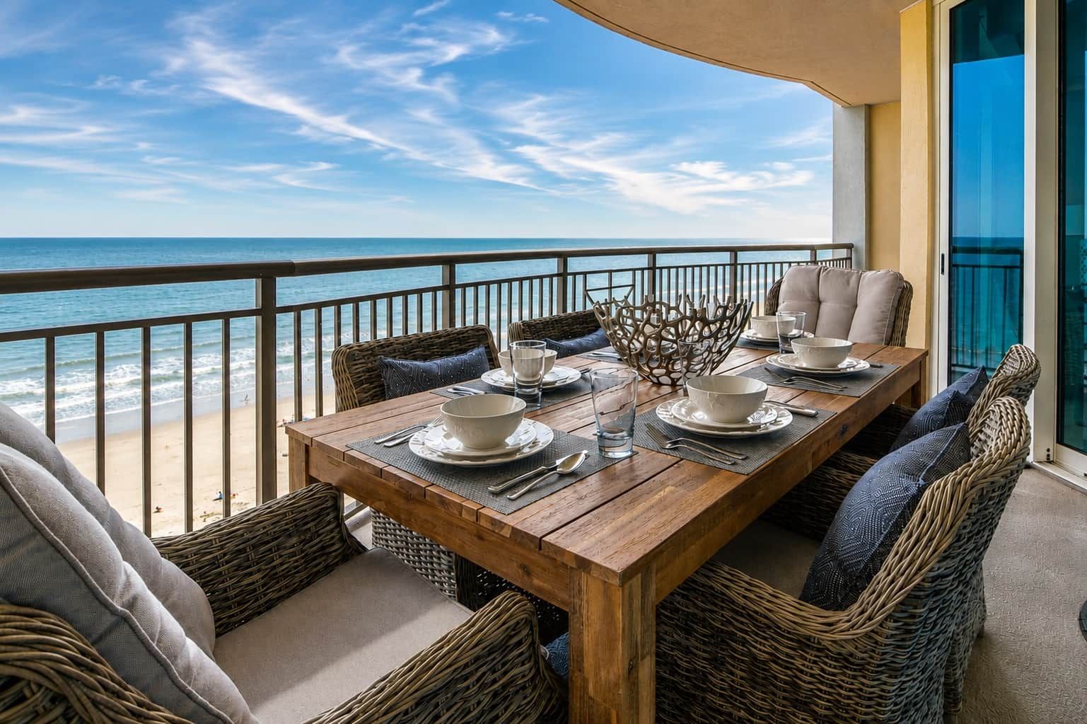 A wooden dining table set for four with white plates and bowls, surrounded by wicker chairs with cushions, overlooking a beach.