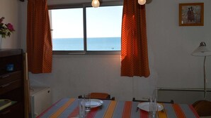 A room with a dining table set for two, a window with curtains, and a view of the sea. - 3-room apt with terrace, AC, parking in Cap d'Agde, near beach and shops (Cap d'Agde)