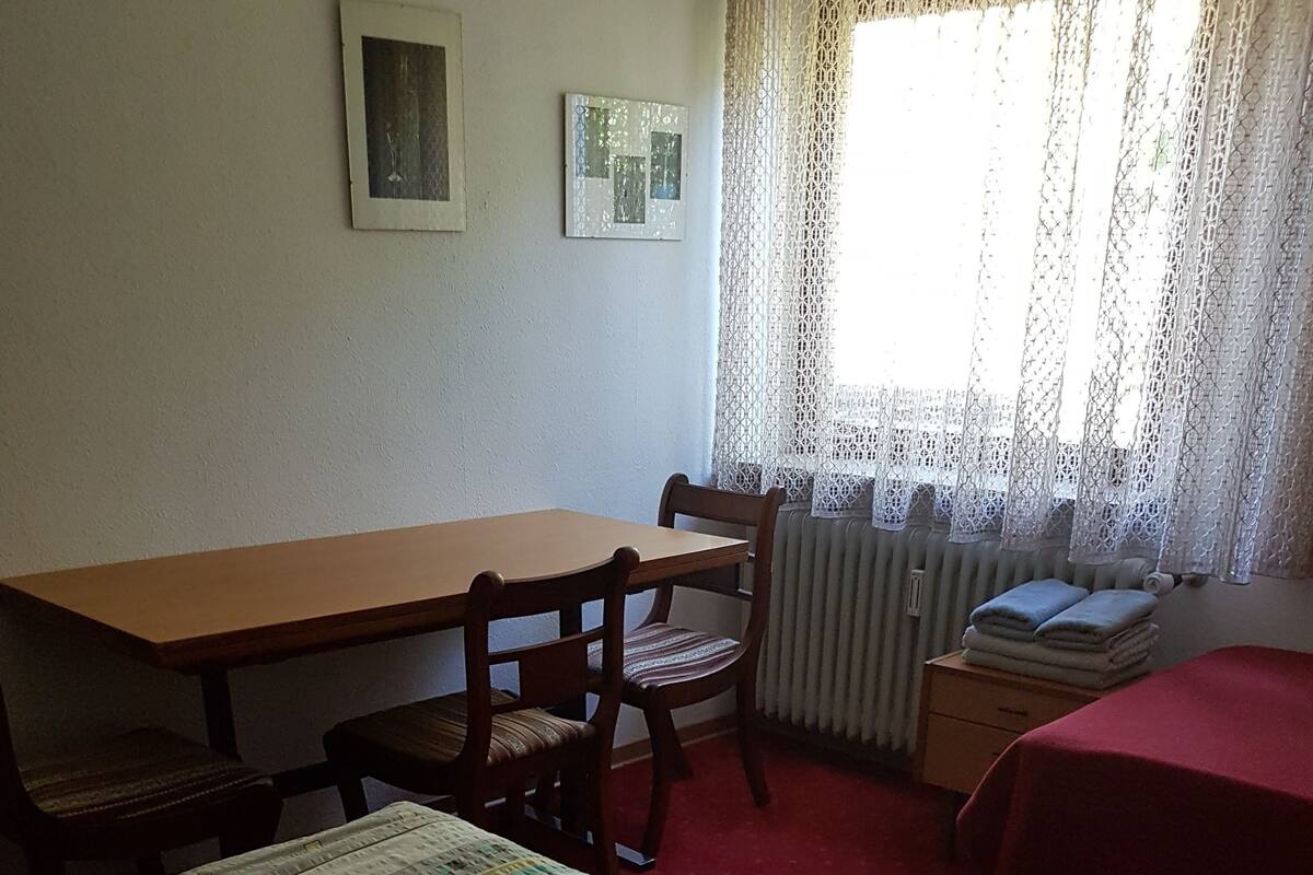 Ein Zimmer mit einem Bett, einem Holztisch, zwei Stühlen, einem Heizkörper und einem Fenster mit Spitzenvorhängen.