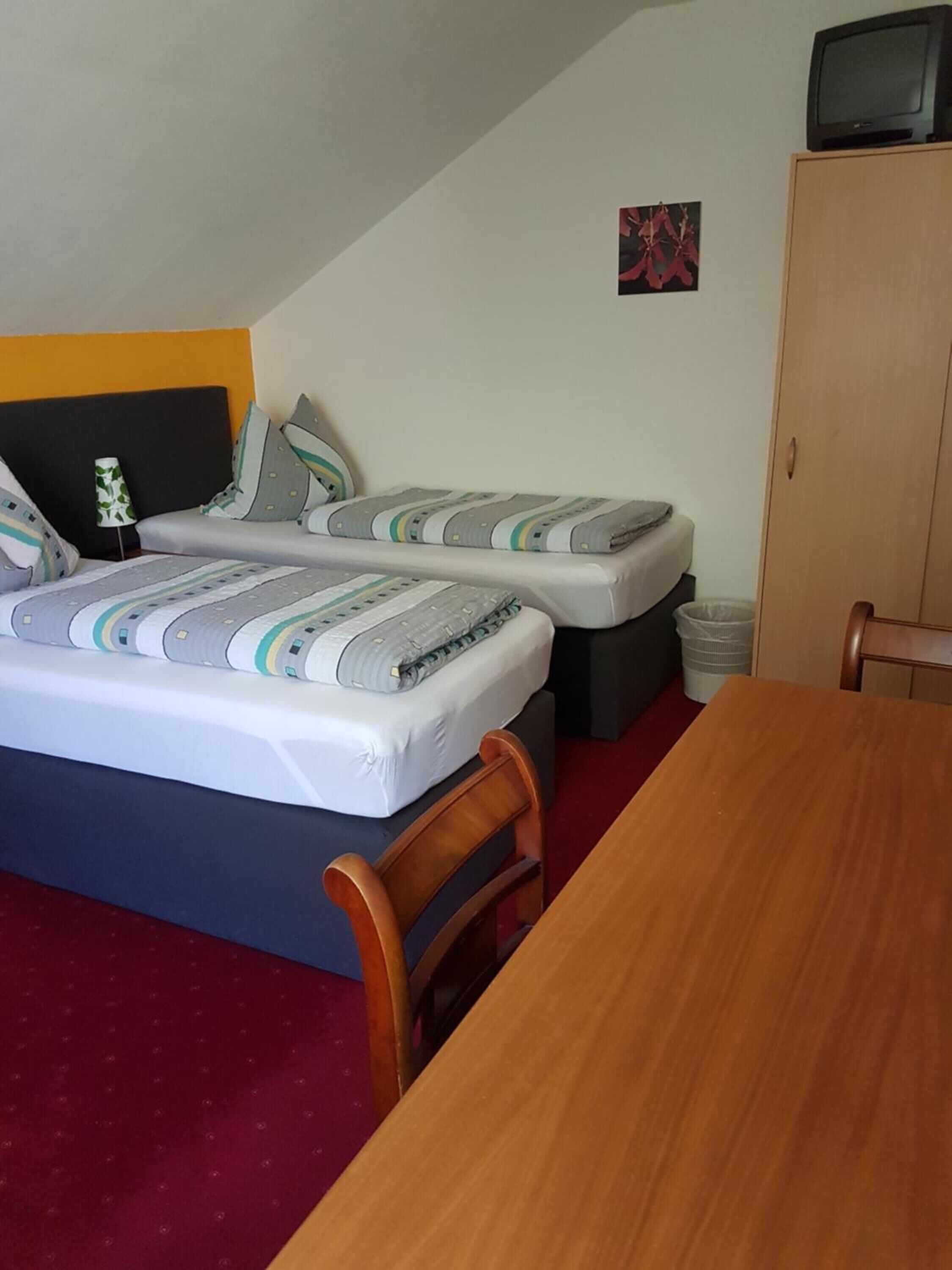 A small, compact room with two single beds, a wooden table, and a television on a cabinet.