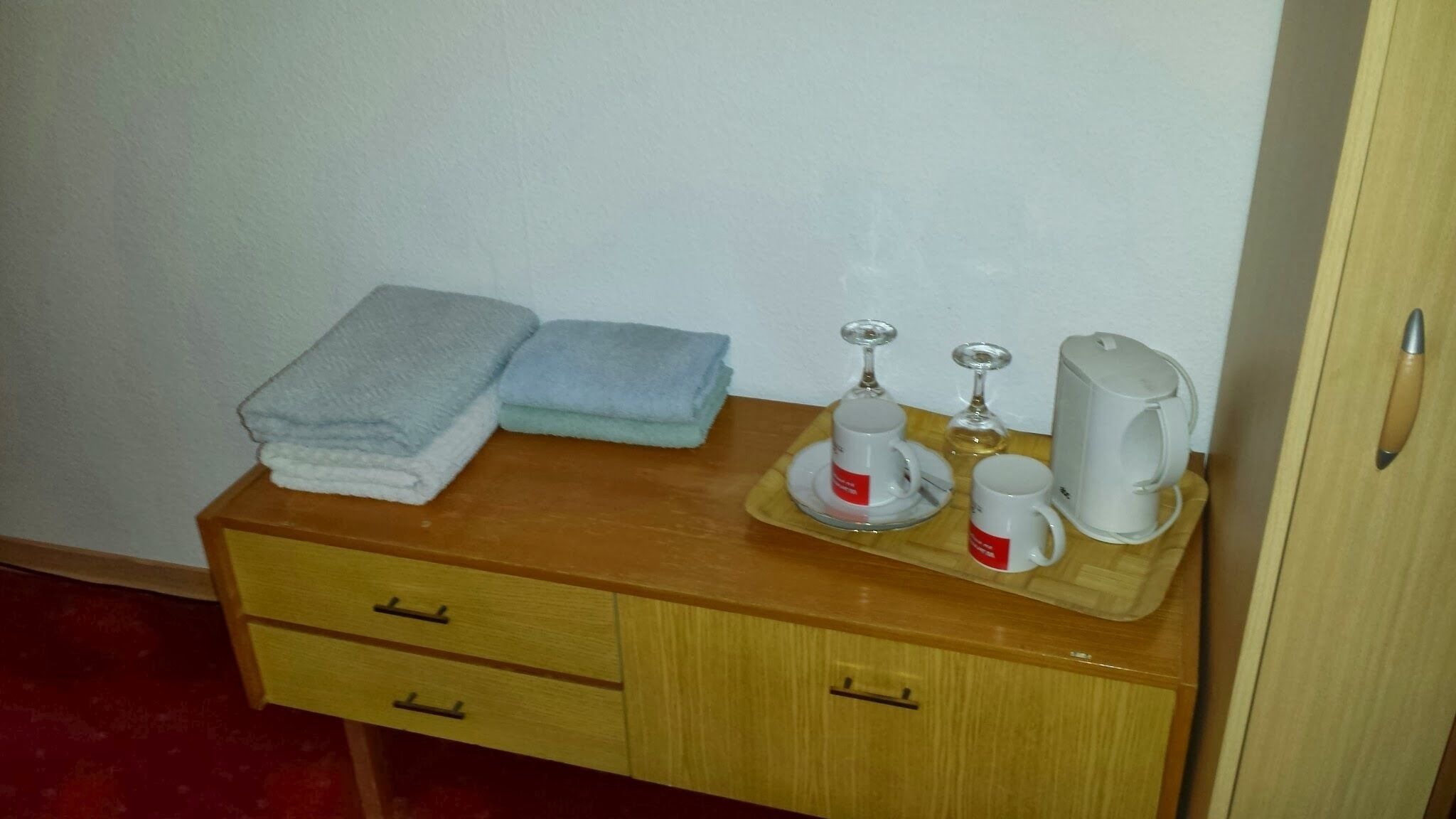 Una mesita de noche de madera con dos toallas, una cafetera, una copa de vino y una taza.