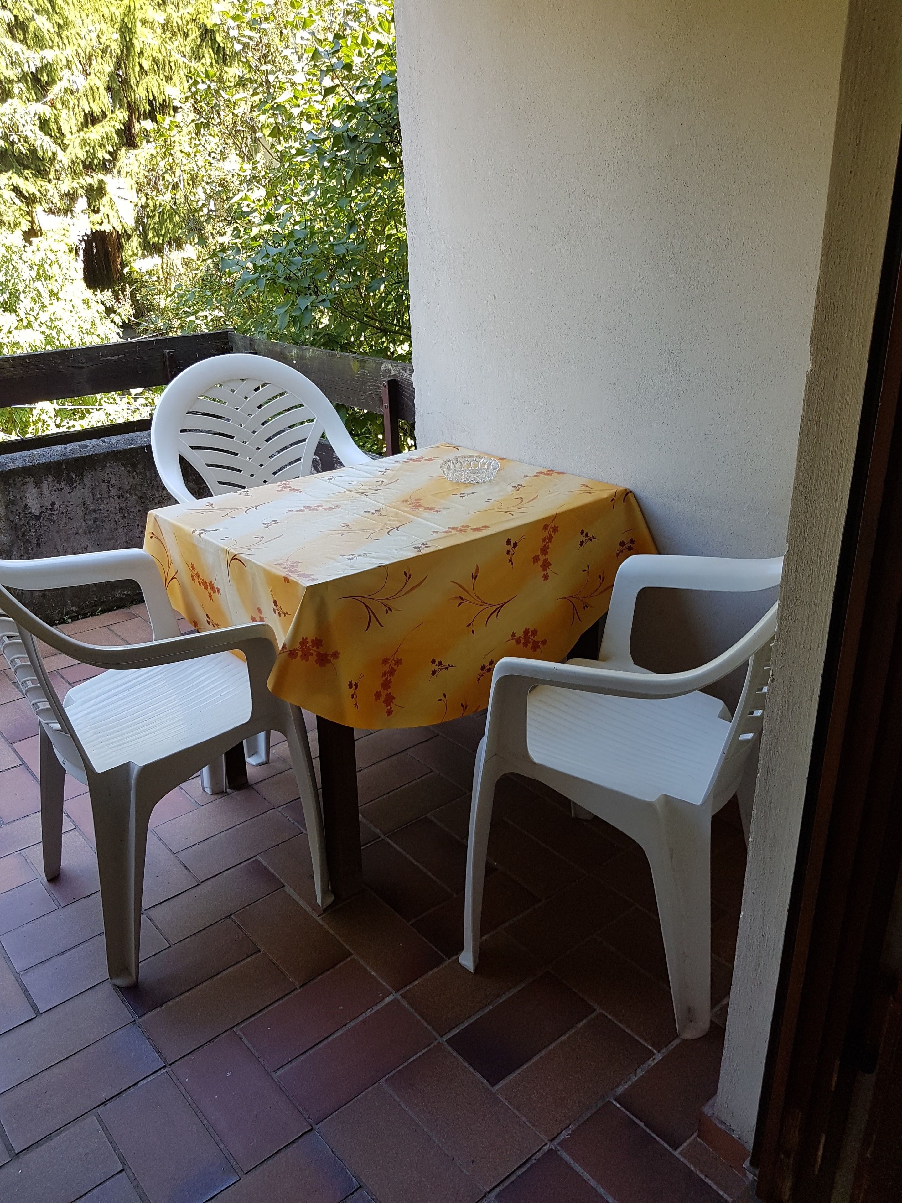 A patio area with a table covered by a floral tablecloth, surrounded by white plastic chairs.