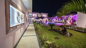A well-lit pathway leading to a modern building with a pool area, outdoor seating, and a garden with tropical plants.