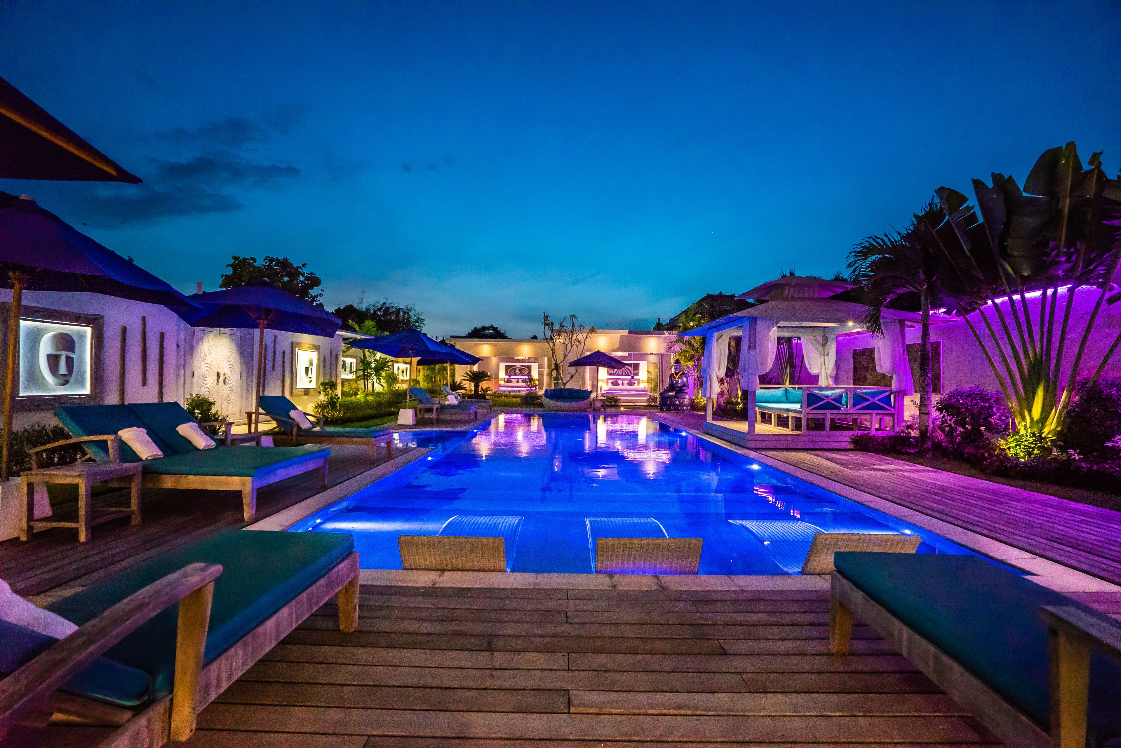 A swimming pool with lounge chairs and umbrellas, surrounded by buildings with illuminated exteriors.