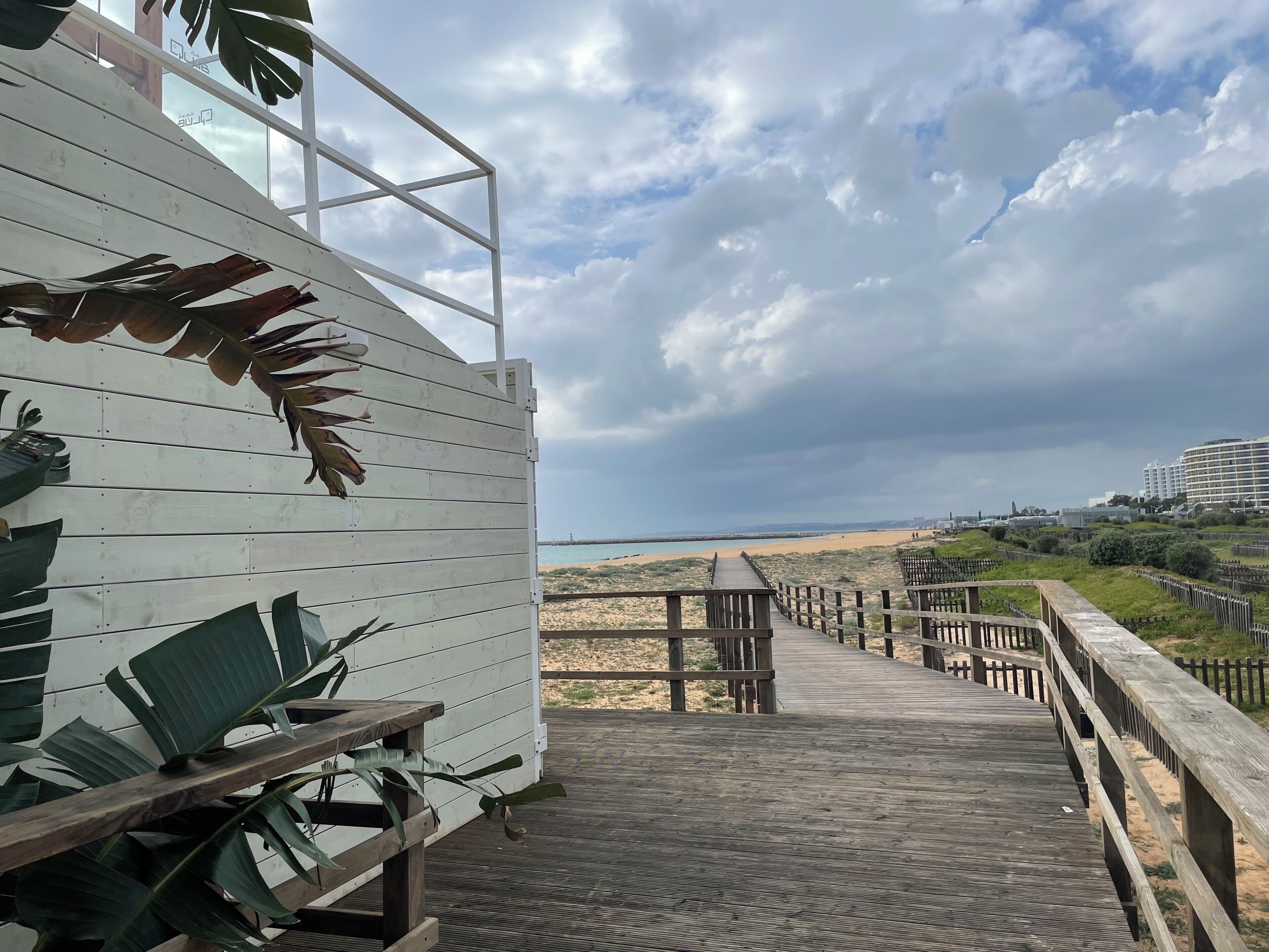 Une terrasse en bois qui mène à une plage, d’où l’on peut admirer l’océan et, au loin, le paysage urbain.