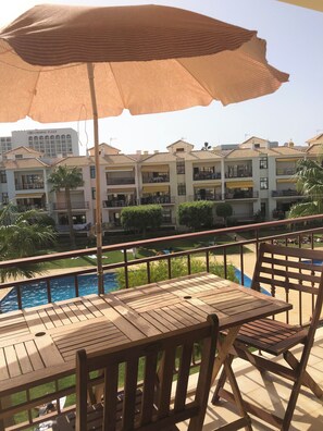Una terraza de madera con una mesa y sillas, con vista a una piscina y un jardín con palmeras.