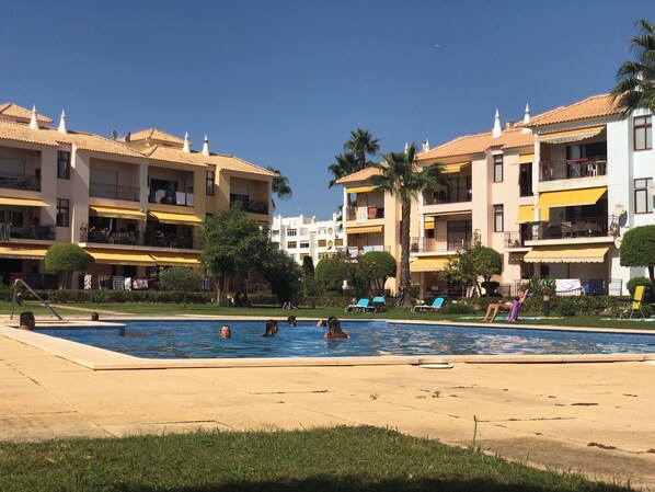 Zona de piscina con varias piscinas, palmeras y un edificio con balcones y mobiliario exterior.