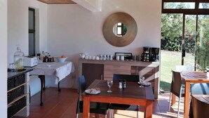 Een moderne keuken met houten balken, een eethoek met een tafel en stoelen, en uitzicht op een tuin door grote ramen.