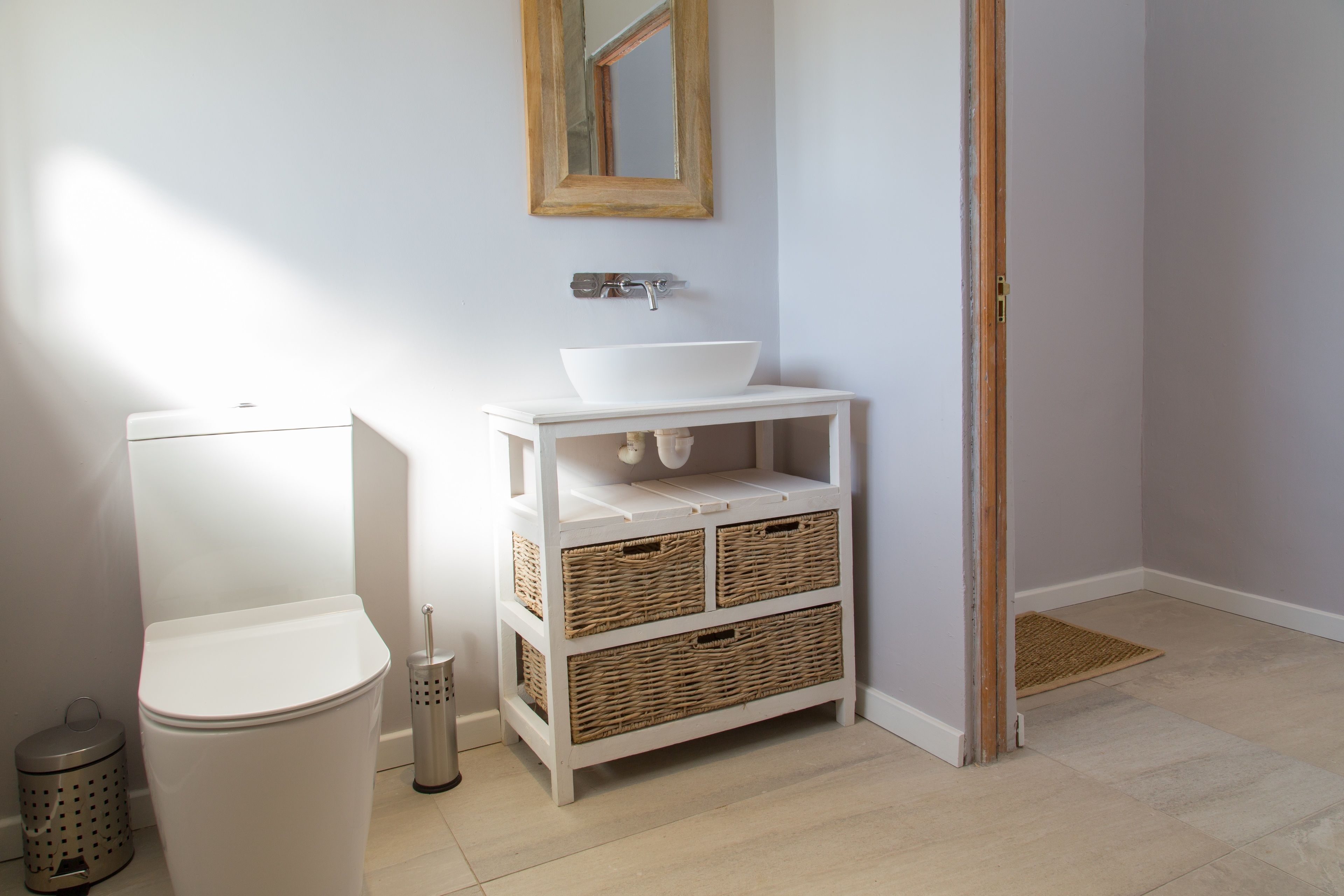 A bathroom with a white toilet, a white sink with a mirror above it, wicker baskets under the sink, and a wooden door.