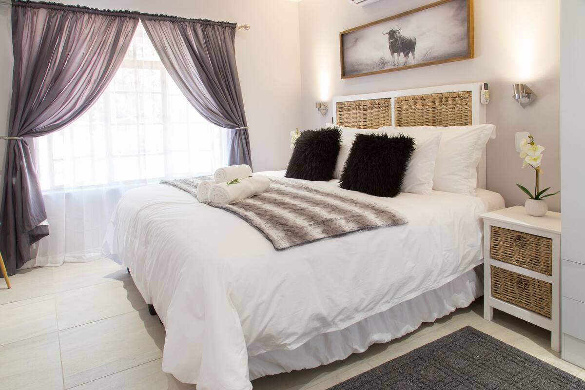 A neatly made bed with white linens, two black pillows, and a striped throw blanket. There is a bedside table with a lamp and a small potted plant. A large window with sheer curtains allows natural light into the room.