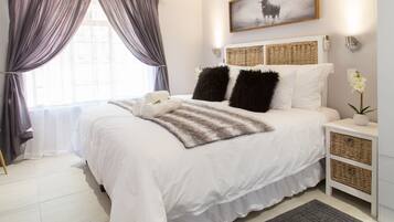 A neatly made bed with white linens, two black pillows, and a striped throw blanket. There is a bedside table with a lamp and a small potted plant. A large window with sheer curtains allows natural light into the room.