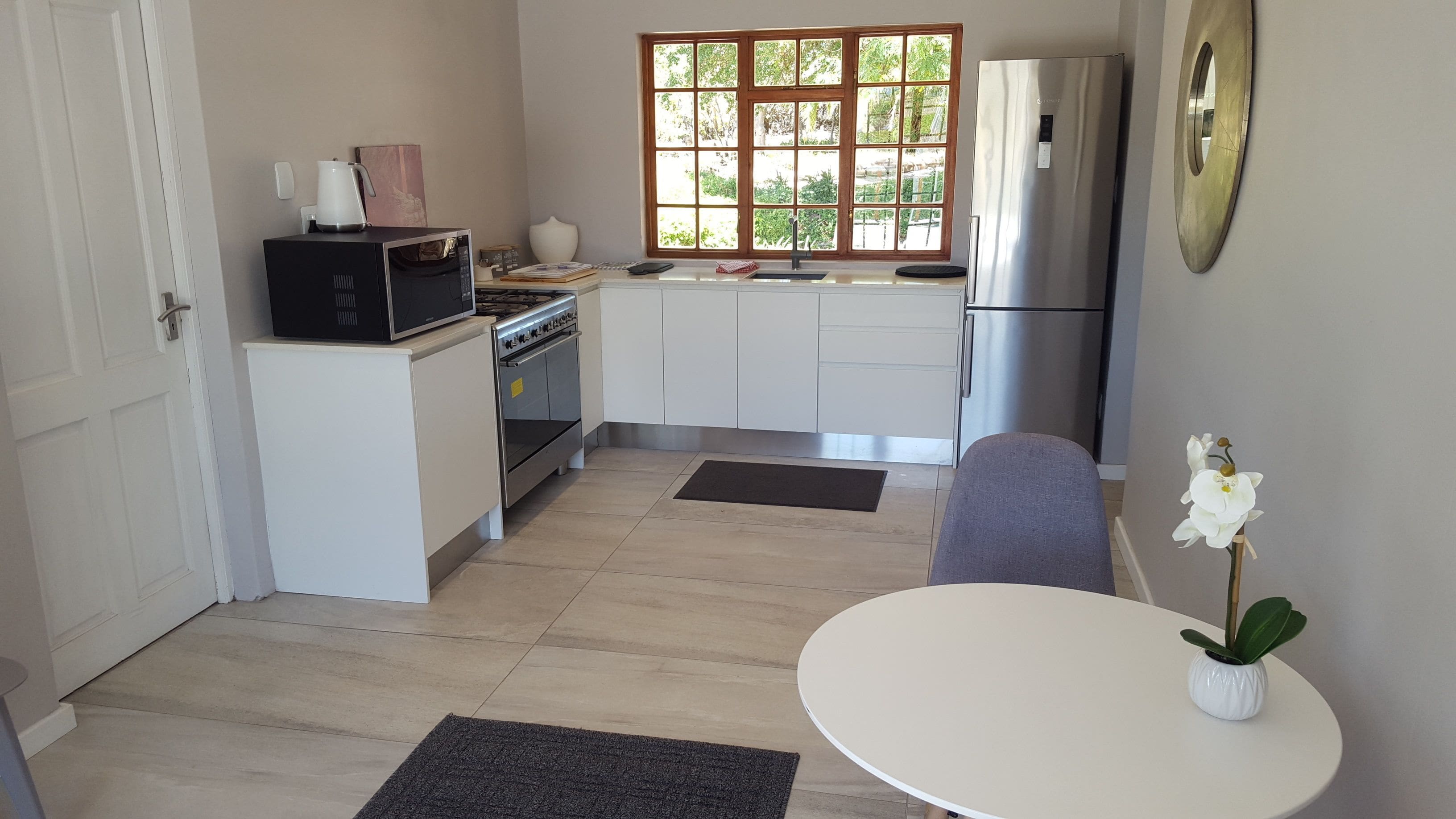 Cocina compacta con microondas, estufa, pileta y refrigerador de acero inoxidable. Hay una ventana con vista a follaje, una mesa blanca con una planta y una puerta a la izquierda.