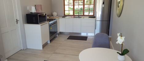 Cocina compacta con microondas, estufa, pileta y refrigerador de acero inoxidable. Hay una ventana con vista a follaje, una mesa blanca con una planta y una puerta a la izquierda.